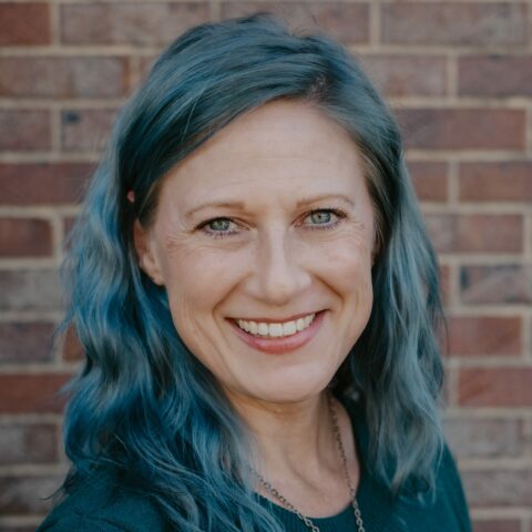 A white woman with blue wavy hair and bright smile stands in front of a brick wall.