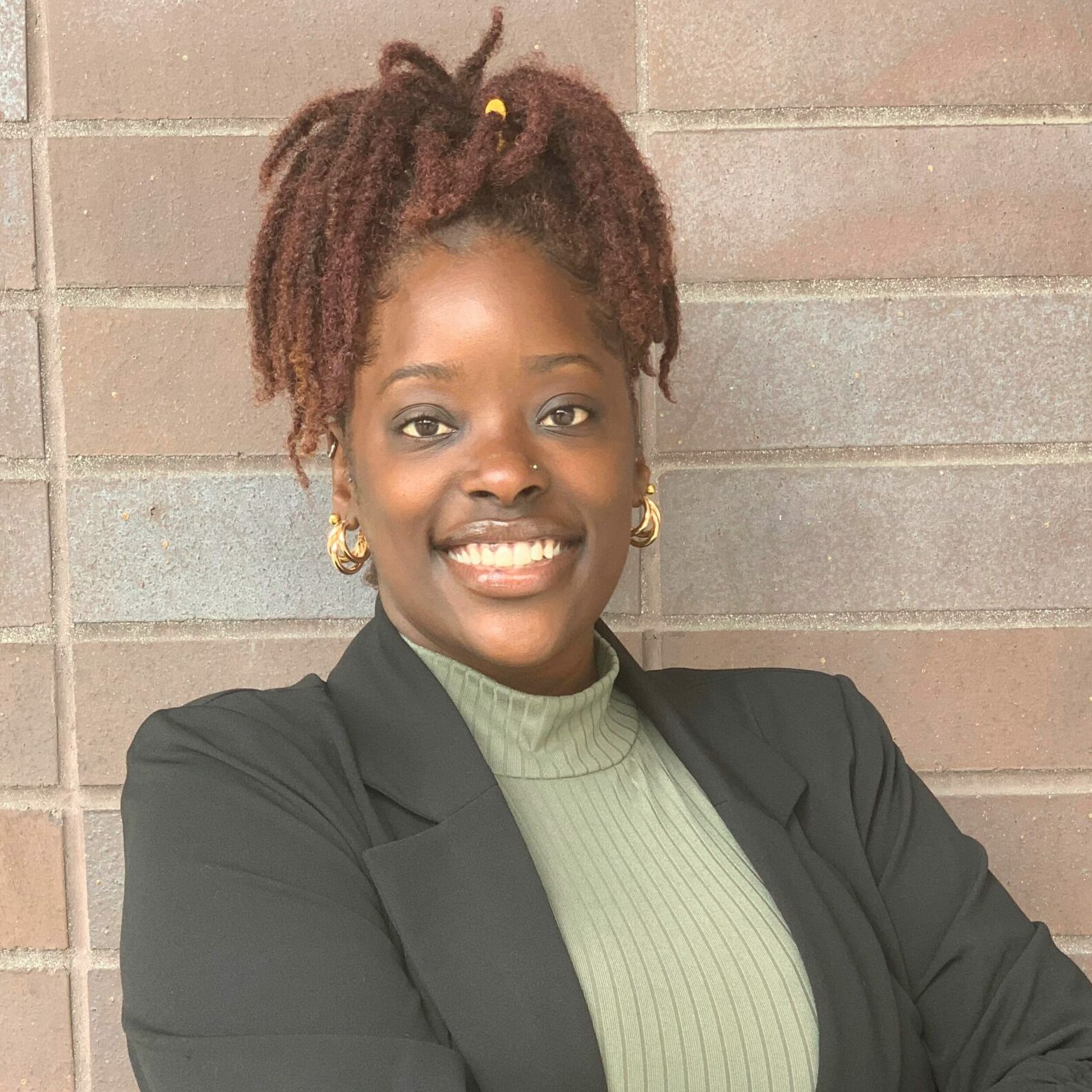 Ashley, a black woman with locs, smiles broadly with her arms crossed over her black suit jacket and green turtleneck.