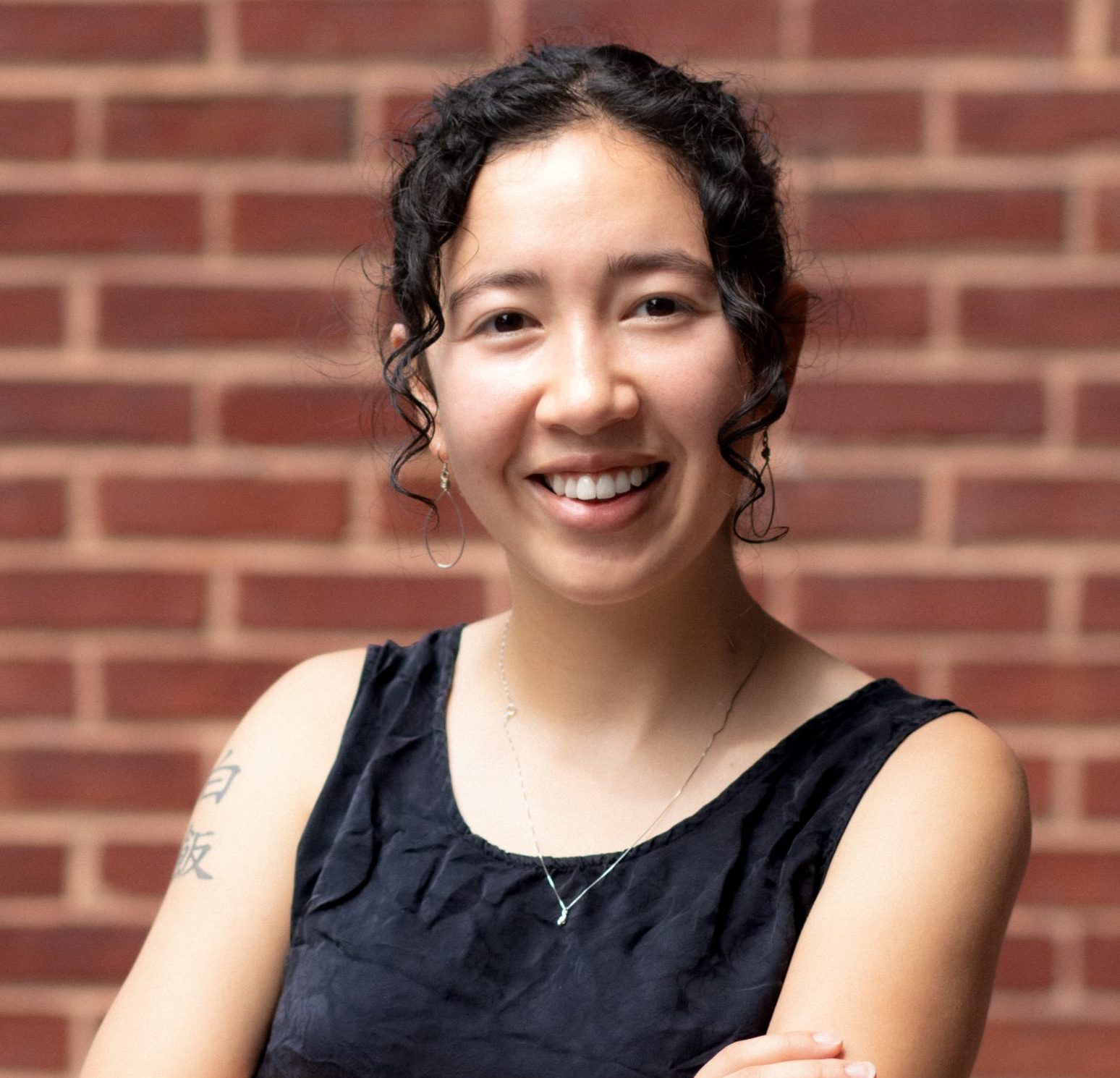 Bianca, a white and Asian woman with curly hair, is smiling and wearing a black top.