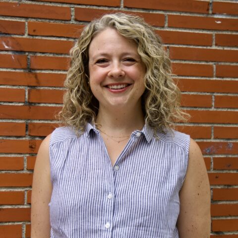 Sarah, a white women with curly blond hair, smiles while wearing a blue and white striped collared shirt.