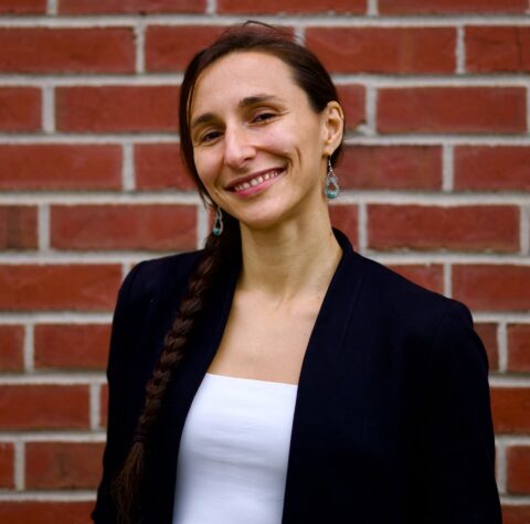 Yoana, a woman with light complexion and long brown braided hair, is smiling while standing in front of a brick wall.