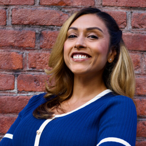 Melanie, a tan Hispanic woman with medium-dark hair displays a warm smile while positioned in front of a brick wall.