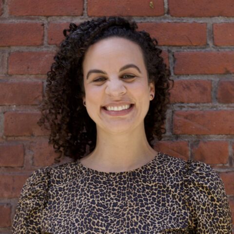 Kelsey, a black and white biracial woman with shoulder length curly hair is wearing a cheetah print shirt and smiling broadly.
