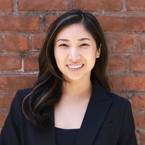 Heile (high-la), a Filipino-American woman with long dark hair and wearing a black blazer, smiles squintingly with teeth against a brick wall.