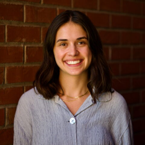 White women with brown hair smiles broadly in a light blue button up.