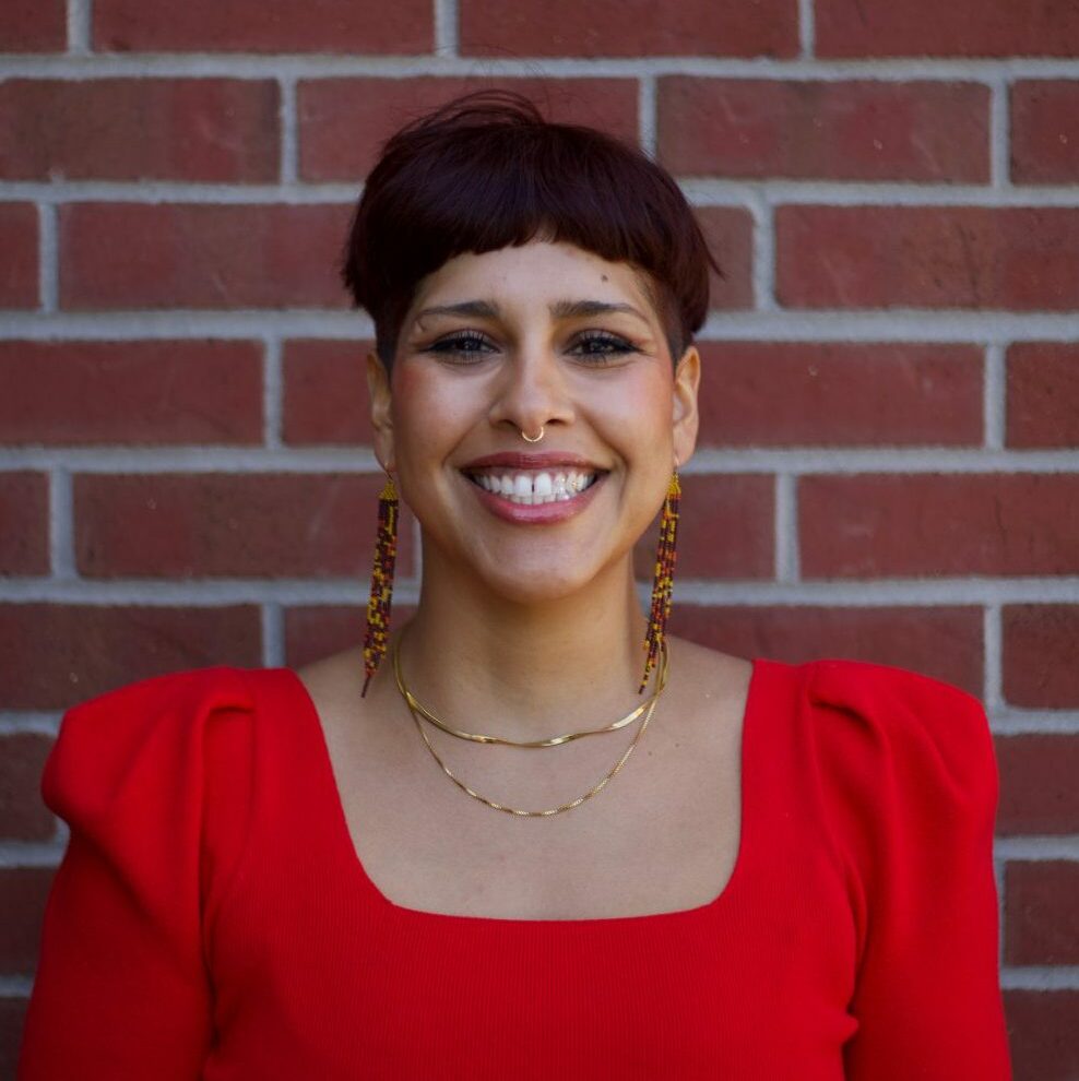 Teresa, a gender nonconforming person with black short hair (pixie cut length) smiles while wearing a red sweater.