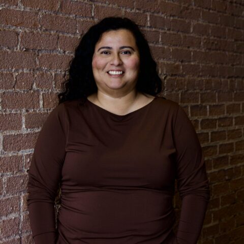 Lesly, a woman with medium complexion and curly dark hair, smiles while wearing a long sleeve, brown shirt.