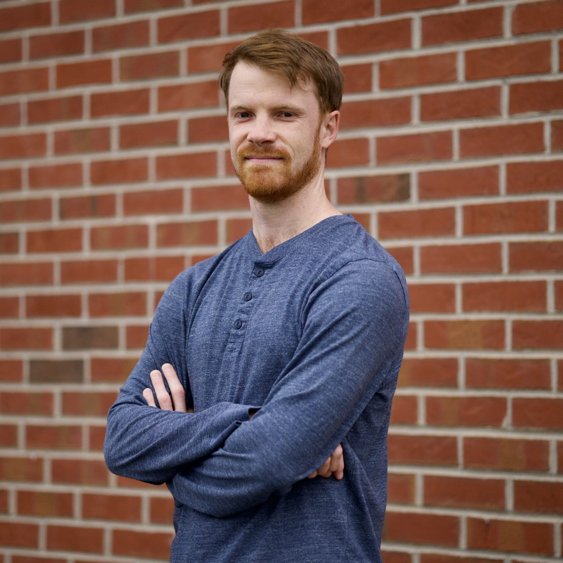 Nicky, a White man with short reddish-brown hair and a beard, stands in front of a brick wall.