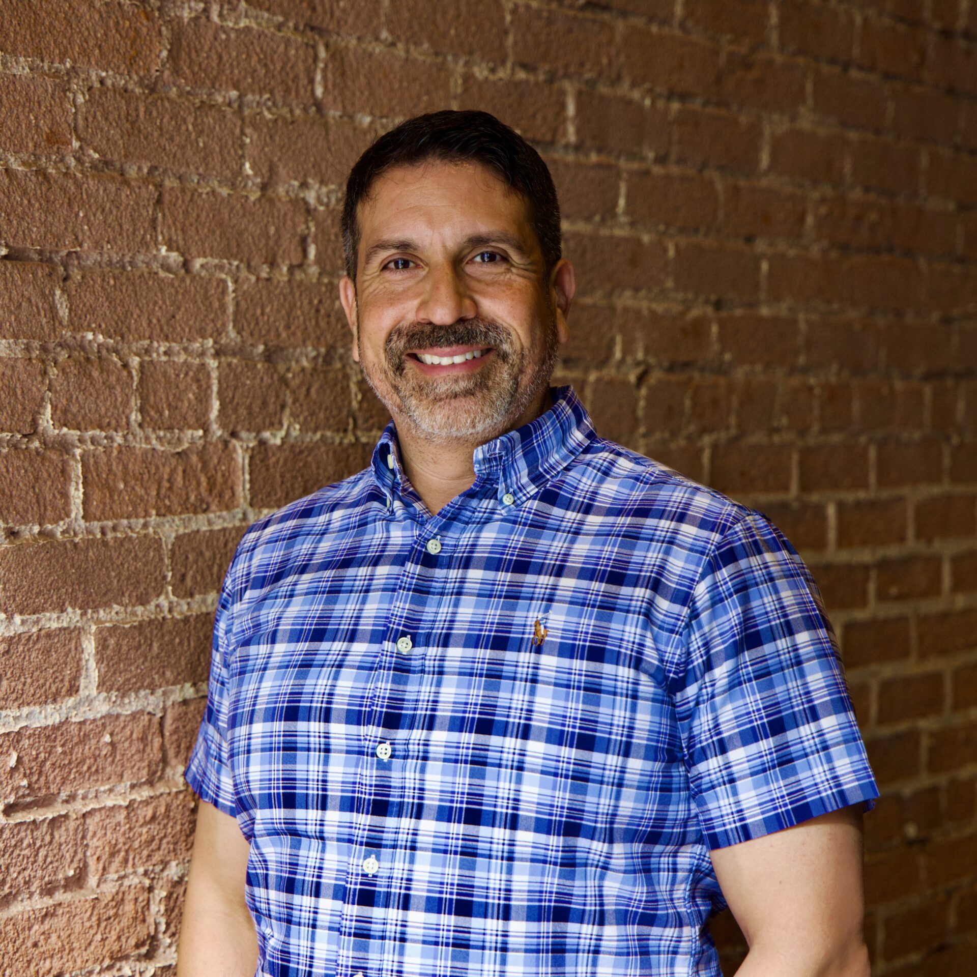Man with beard and wearing a blue plaid shirt, standing in front of a brick wall.
