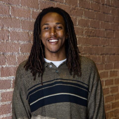 Smiling person with long dreadlocks wearing a striped sweater, standing in front of a brick wall.