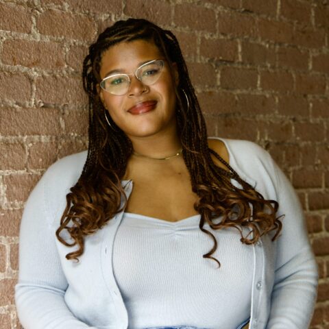 Maya, a Black woman with braids and wearing a blue sweater, smiles while leans against a brick wall.