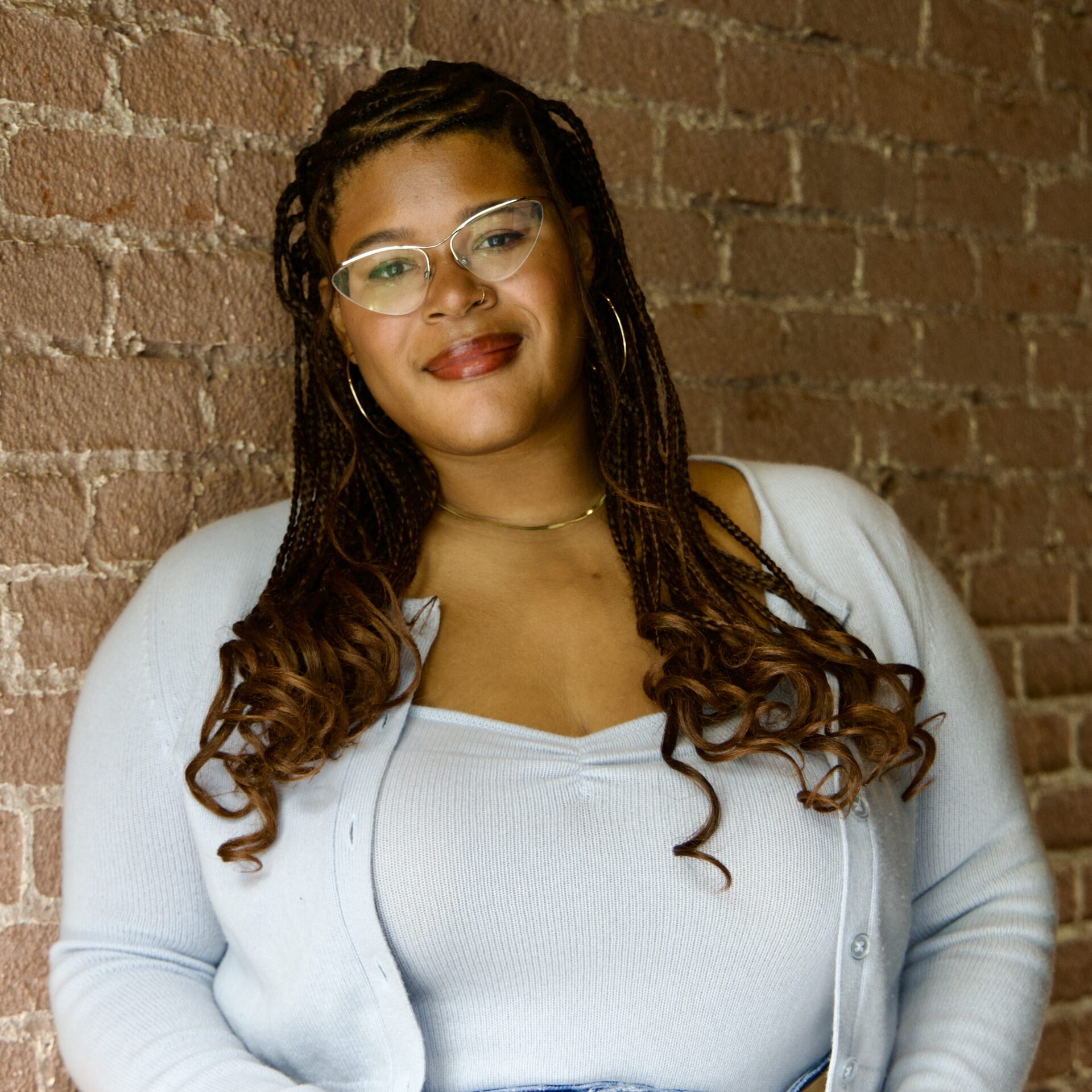 Maya, a Black woman with braids and wearing a blue sweater, smiles while leans against a brick wall.