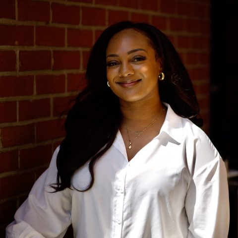 Ashaki Scott, a Black and Japanese woman with long dark hair, is smiling warmly in a white blouse and gold jewelry, standing against a red brick wall with sunlight on her face.