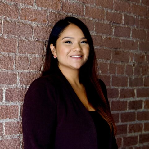 A smiling woman with long dark hair stands in front of a brick wall.