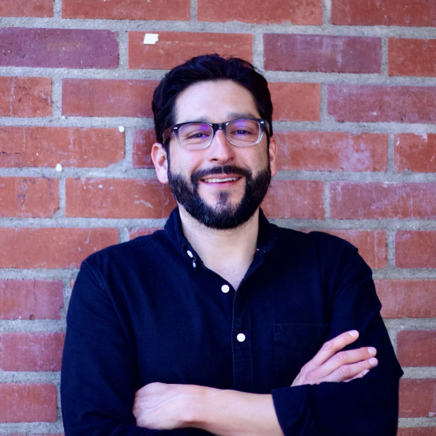 Man with dark hair, glasses, and black shirt standing against a brick wall.