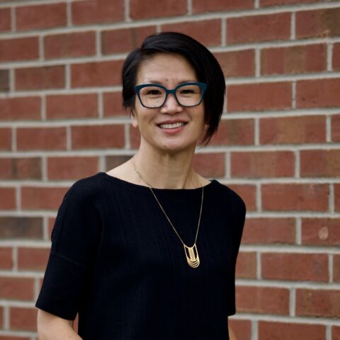 Alison, Asian woman smiling. She has black hair with a short, asymmetrical haircut, turquoise glasses, wearing a black top.