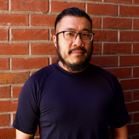 Man in glasses, in black standing in front of a brick wall.