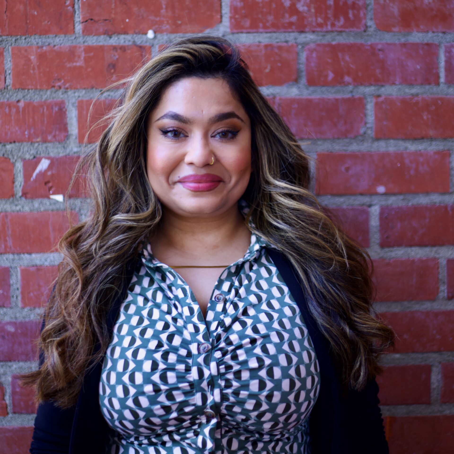 Rabia, a Middle-Eastern woman with long brown highlighted hair, wears a nose ring and printed shirt in front of a brick wall.