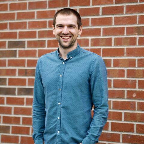 A white man in a green button-up shirt smiles in front of a brick wall.