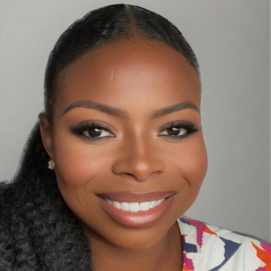 LaShaye, a Black woman with black hair in a side ponytail over her shoulder, smiles broadly while wearing a colorful printed shirt.