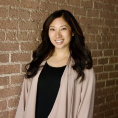 Heile (high-la) Gantan-Keo, a Filipino-American woman with long dark hair, smiles in front of a brick wall.