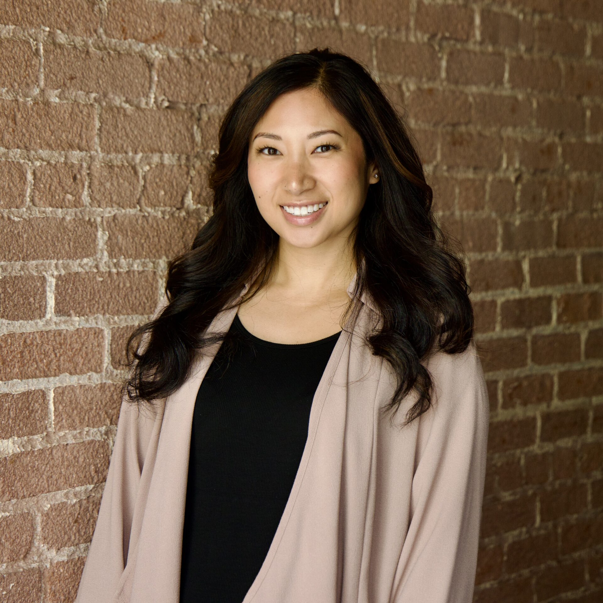 Heile (high-la) Gantan-Keo, a Filipino-American woman with long dark hair, smiles in front of a brick wall.