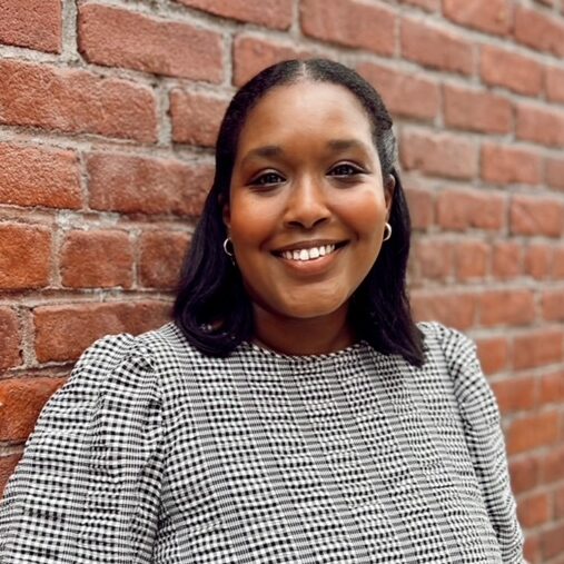 Hawa, a black woman with shoulder length black hair is smiling against a brick wall.