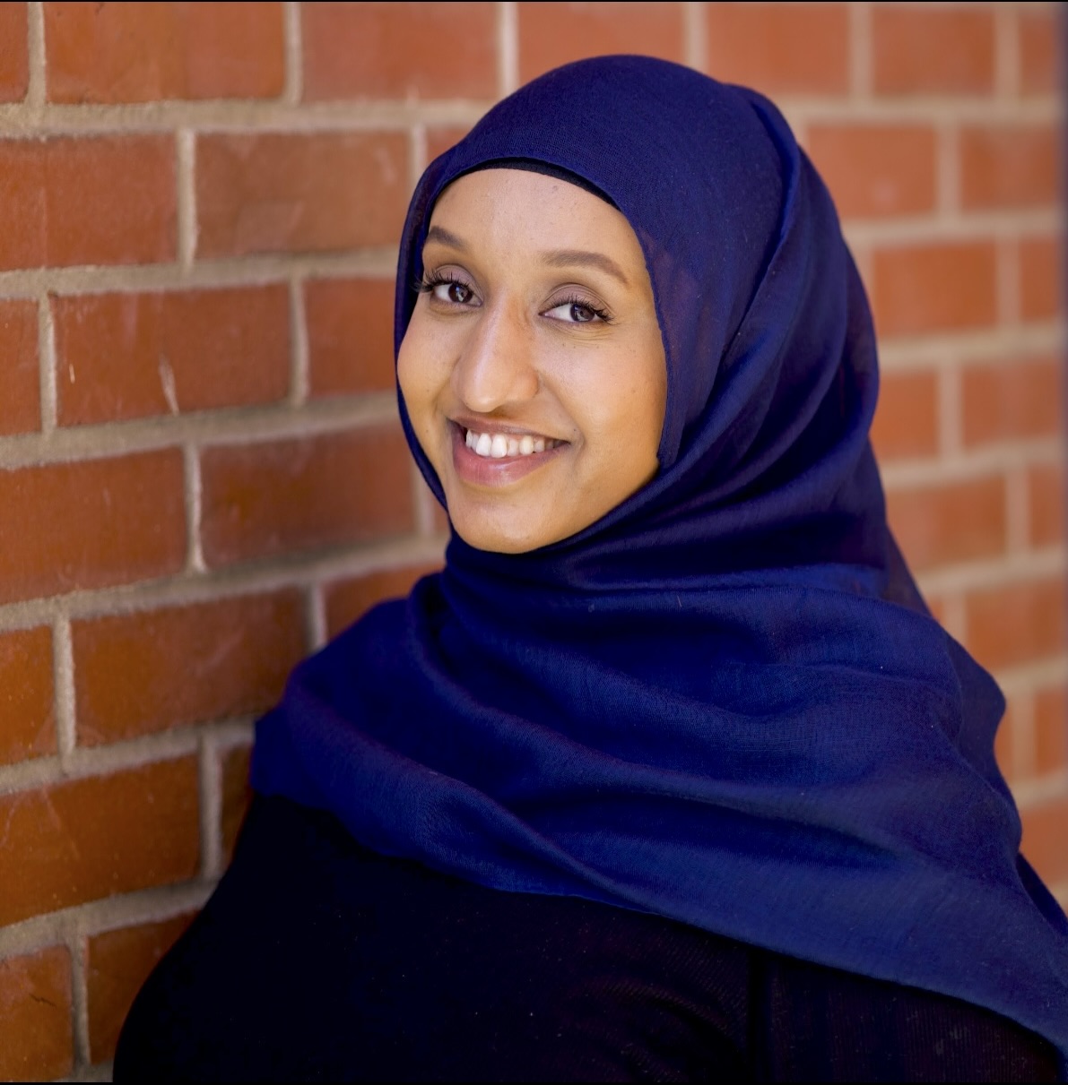 A woman in a navy blue hijab smiling in front of a brick wall.