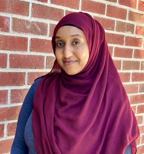 A woman in a maroon hijab stands against a brick wall, smiling softly with her mouth closed.