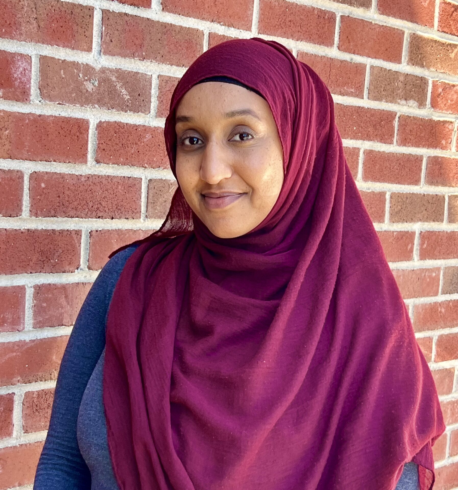 A woman in a maroon hijab stands against a brick wall, smiling softly with her mouth closed.