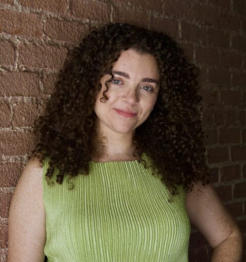 A woman wearing a green shirt stands against a brick wall. She has curly hair that reaches just past her shoulders.