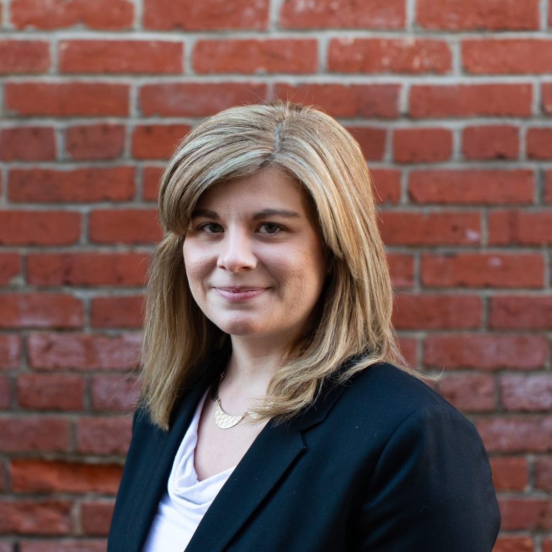 Kayla, a while woman with shoulder-length dirty blonde hair, wearing a white shirt under a black blazer, stands in front of a brick wall.