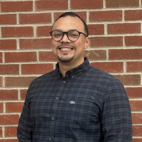 Man wearing glasses and a dark plaid button-down shirt, smiling in front of a red brick wall.