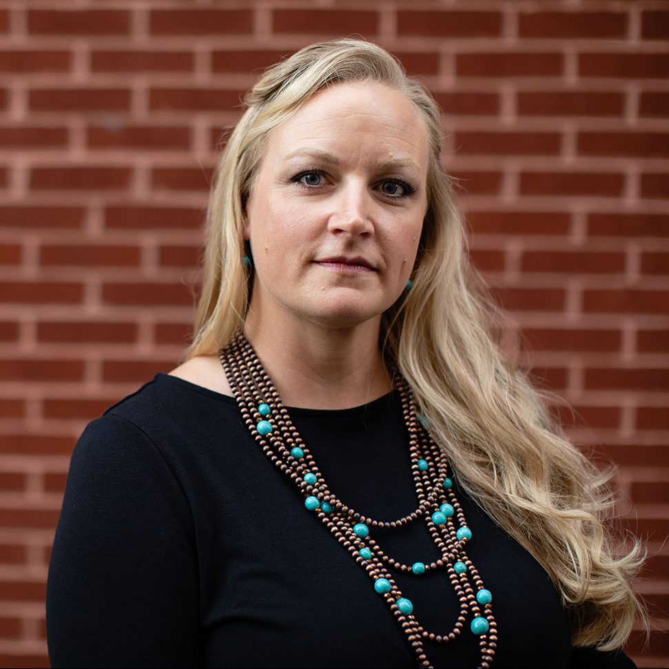 Talia is a white woman with long blonde hair, wearing a slight smile, black shirt, and long turquoise and bronze necklace.