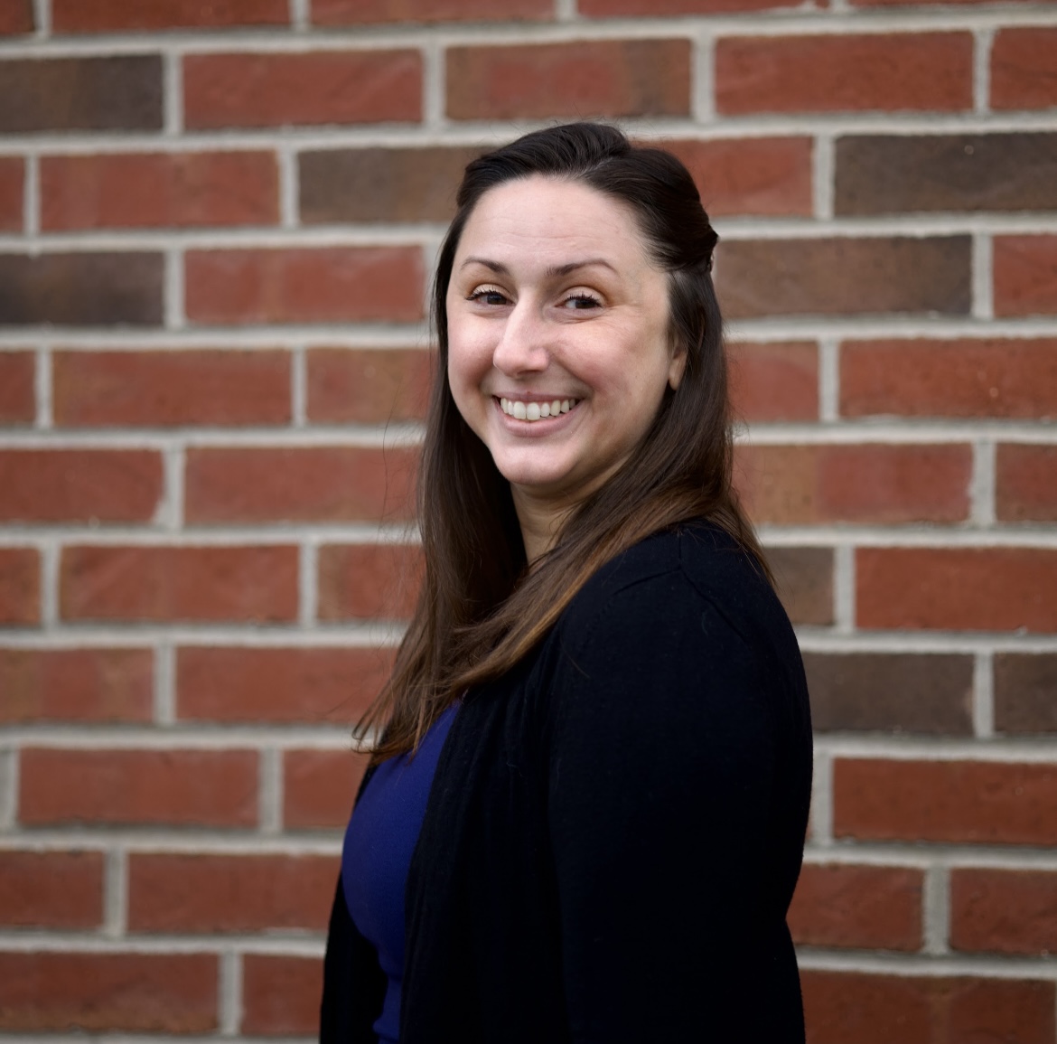 Megan, a woman with brown hair pinned half up, is standing in front of a brick wall wearing a blue top and black sweater.