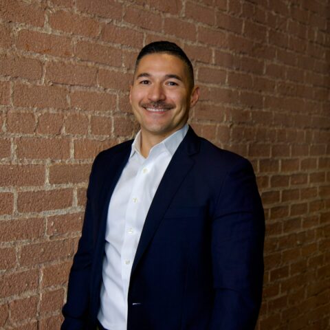 A man in a suit stands confidently in front of a textured brick wall.
