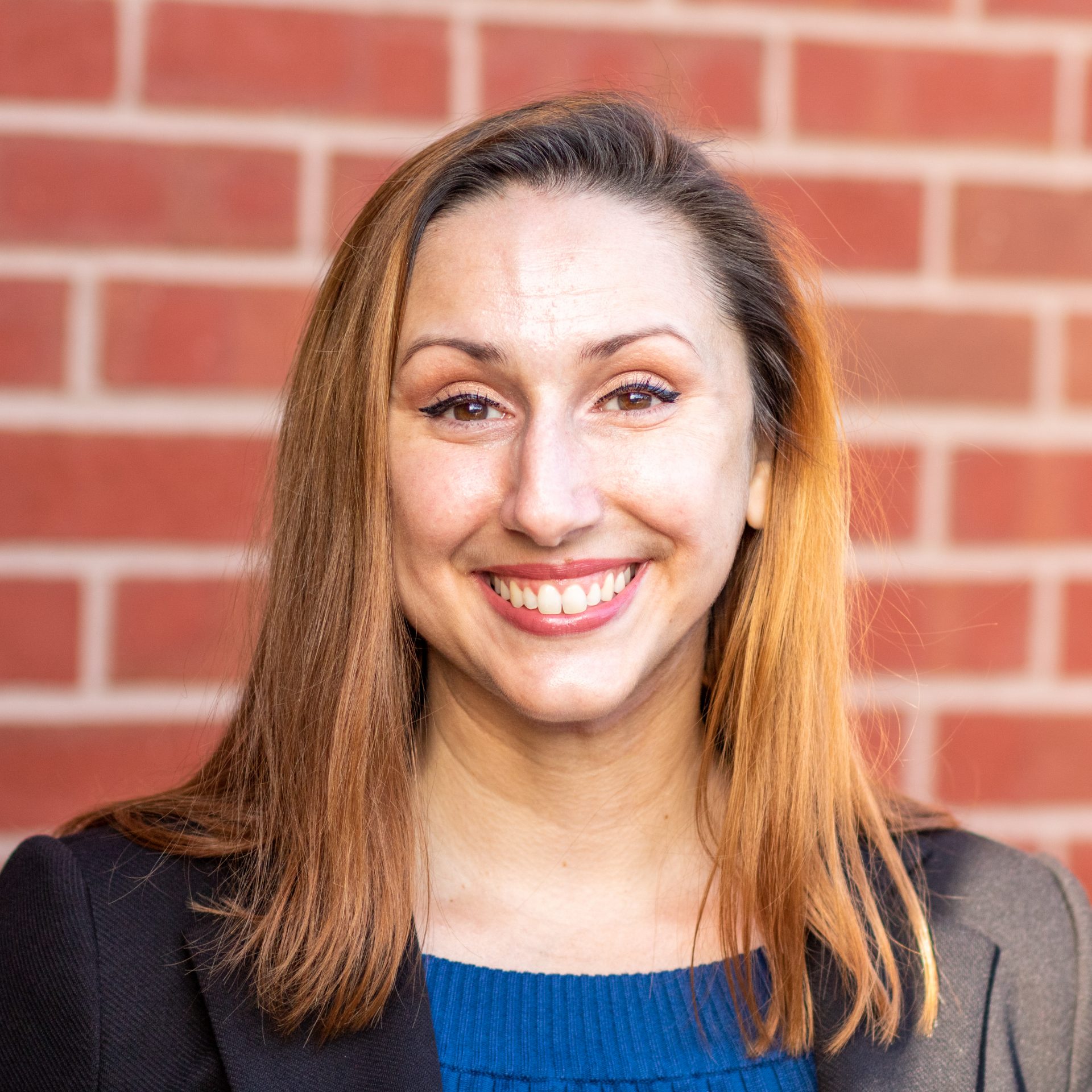 Megan, a woman with a light complexion and light brownish-red hair, is wearing a black blazer over a blue sweater while smiling at the camera.