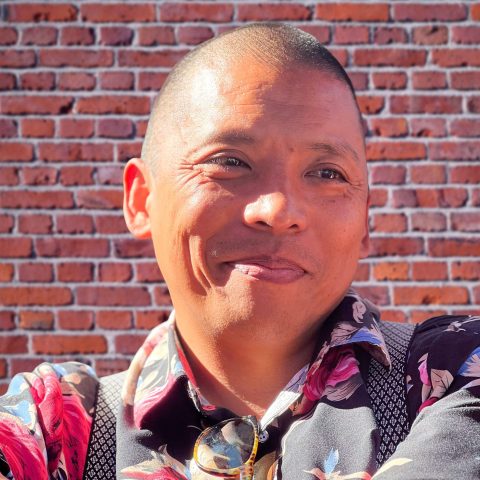 Joey, man dressed in a floral shirt and suspenders stands in front of a textured brick wall.