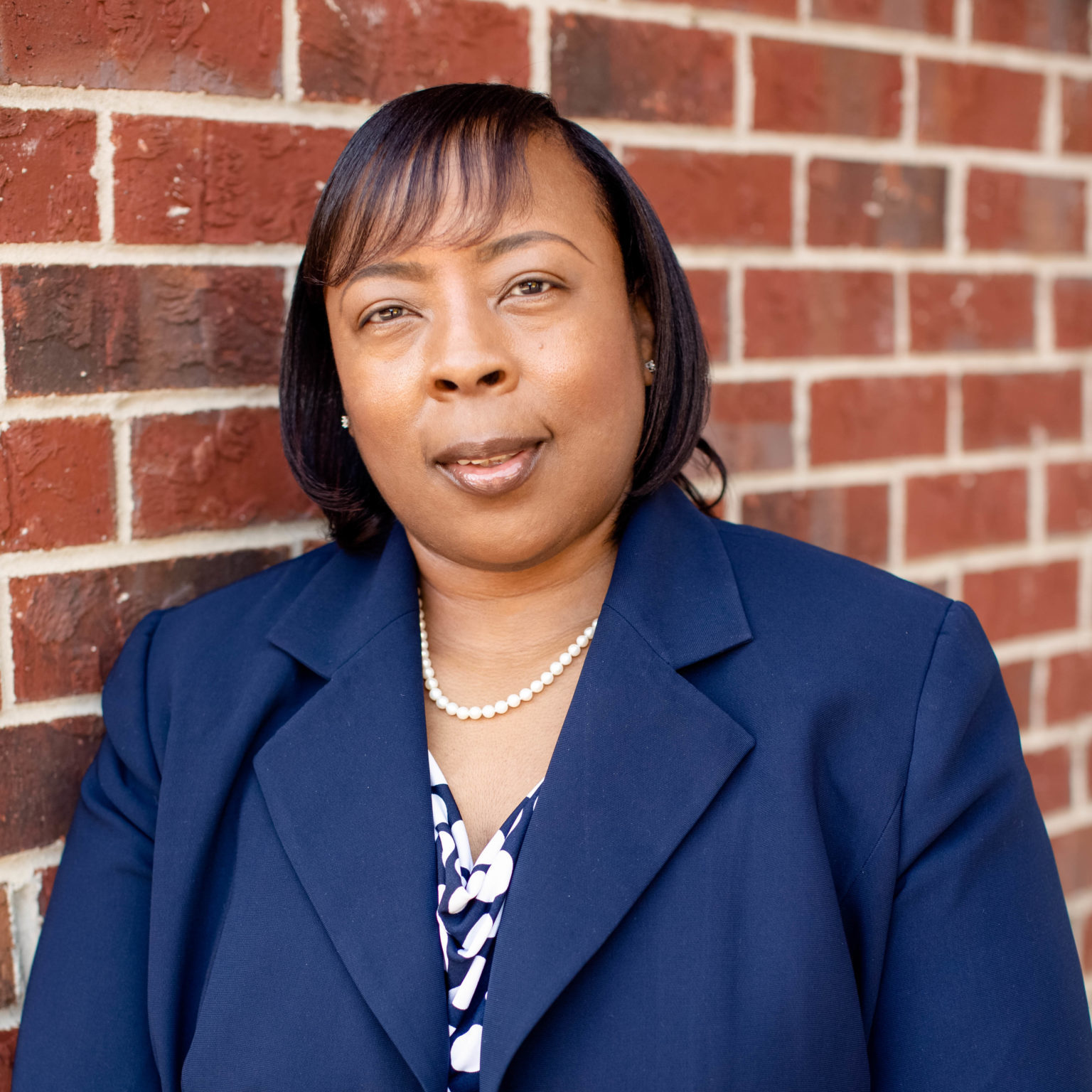 Phebia, an African American woman with shoulder-length brown hair, smiles while wearing a blue jacket and blue and white polka dot shirt.