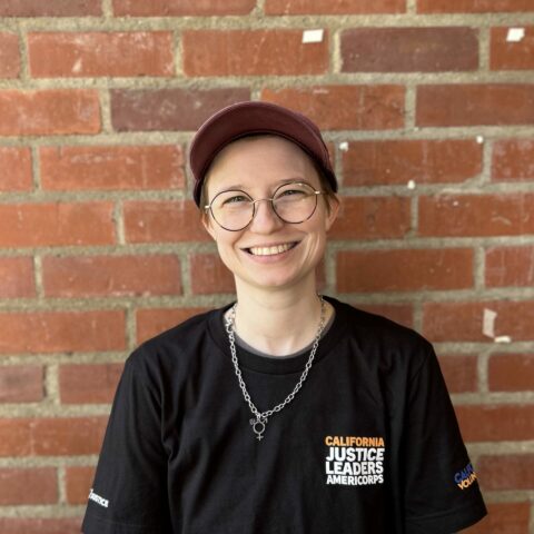 A masculine presenting individual smiling at the camera, with short hair who is wearing a hat and glasses against a brick wall.