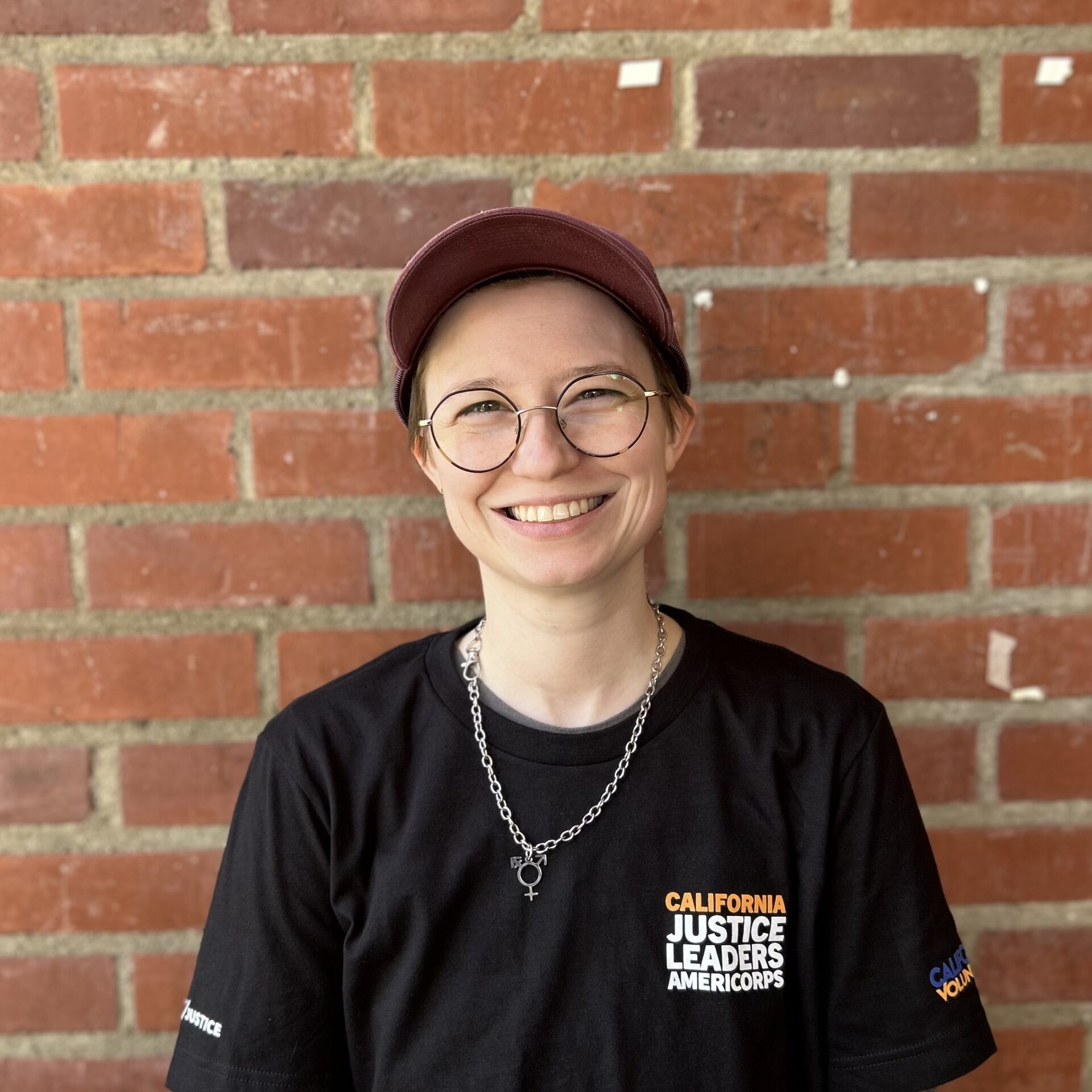 A masculine presenting individual smiling at the camera, with short hair who is wearing a hat and glasses against a brick wall.