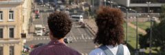 Two young people with dark curly hair, seen from behind, look out at a cityscape from above.