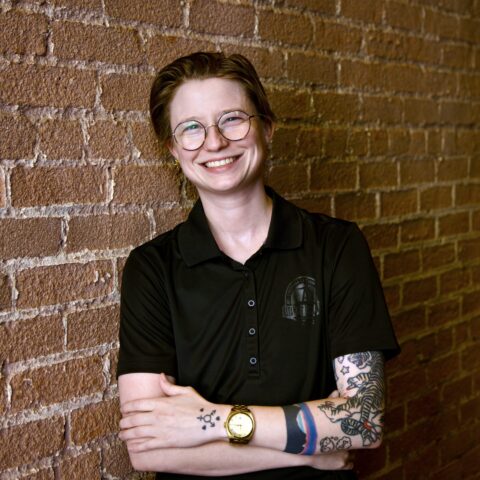 Trans-masculine individual smiling at the camera in a black polo shirt with their arms crossed in front of them. Rob is wearing glasses, has tattoos, and a gold watch.