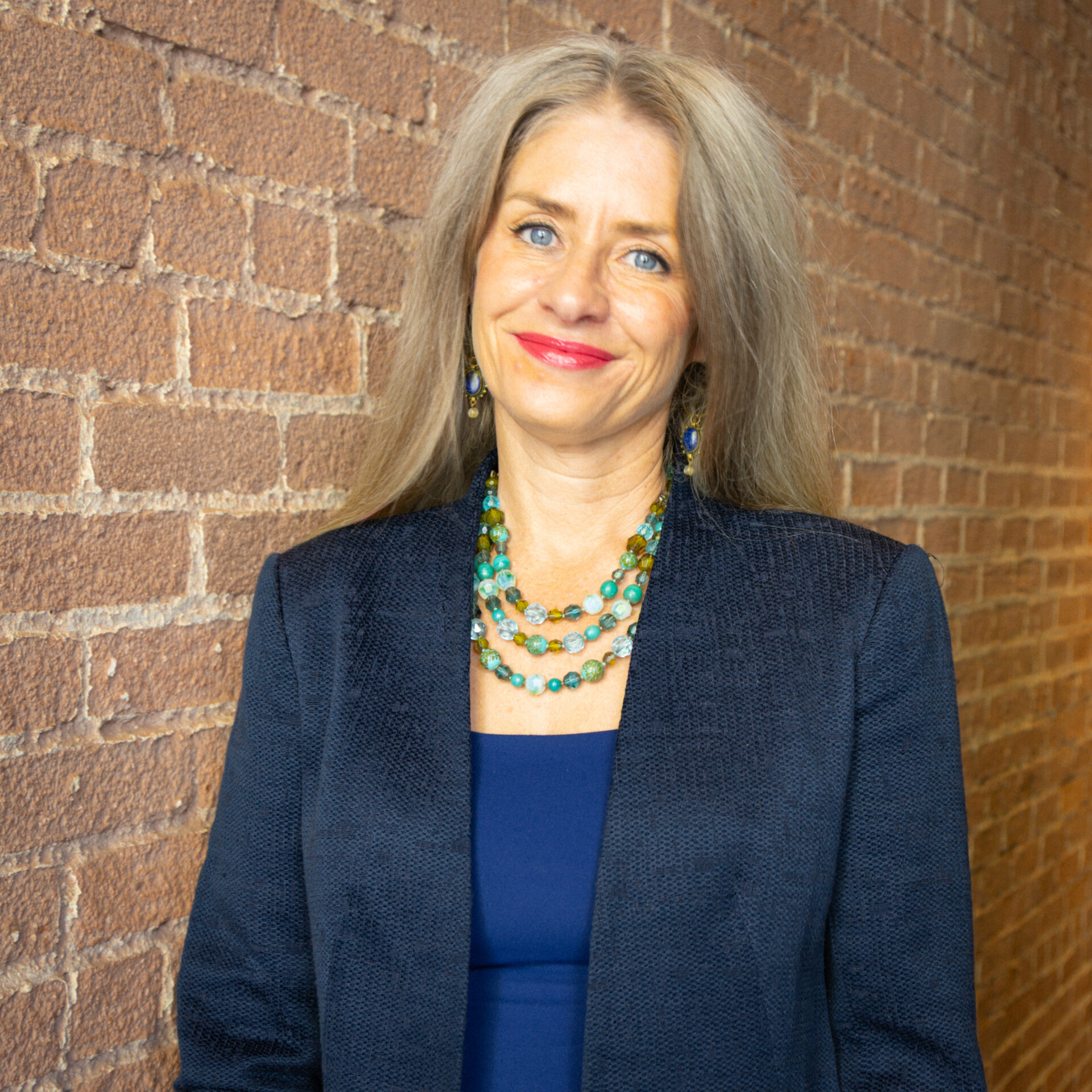 Heather, a woman with long gray hair, a blue blazer, and a colorful necklace, stands smiling against a brick wall.