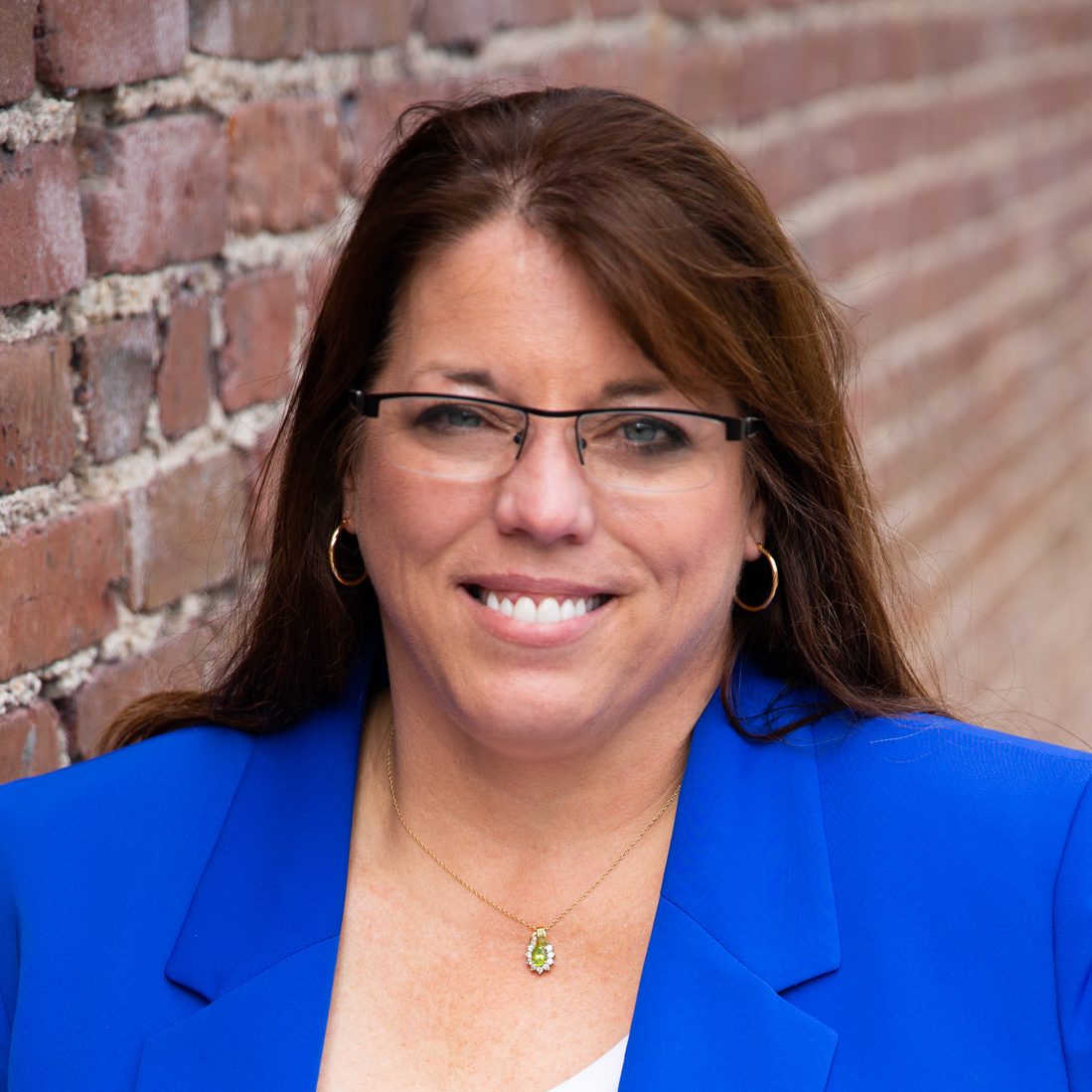 Stroud, a white woman with shoulder-length brown hair, smiles while wearing a blue blazer and black glasses.